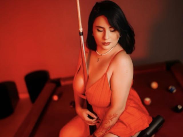 Sara Guzman, con capelli scuri a metà schiena, indossa un vestito rosso, tiene un bastone da biliardo, è inginocchiata, illuminazione rossa, con tatuaggi. braccio, palle da biliardo sul pavimento, posa sexy e seducente, sguardo intenso.