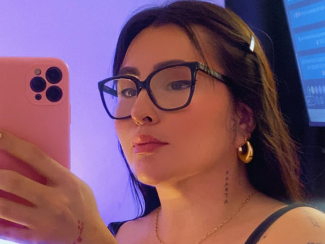 Nahaufnahme-Selfie von SaraGuzman, einer attraktiven Frau mit langen braunen Haaren, schwarzen Brillen, goldenen Creolen und einem schwarzen Tanktop. oben, mit einem rosa iPhone in der Hand. Sie hat einen verführerischen Ausdruck..