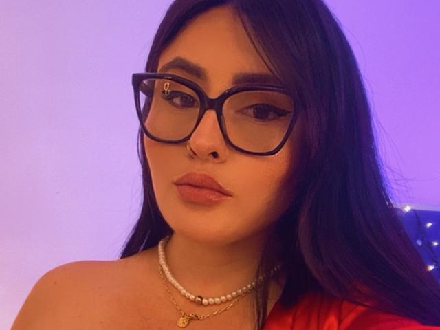 Nahaufnahme von SaraGuzman mit langen, schwarzen Haaren, die eine Brille mit schwarzem Rahmen, eine Perlenkette und ein rotes Oberteil trägt. Ihr Ausdruck ist verführerisch, mit deutlich sichtbarer sexueller Spannung. Der Hintergrund ist blau beleuchtet..