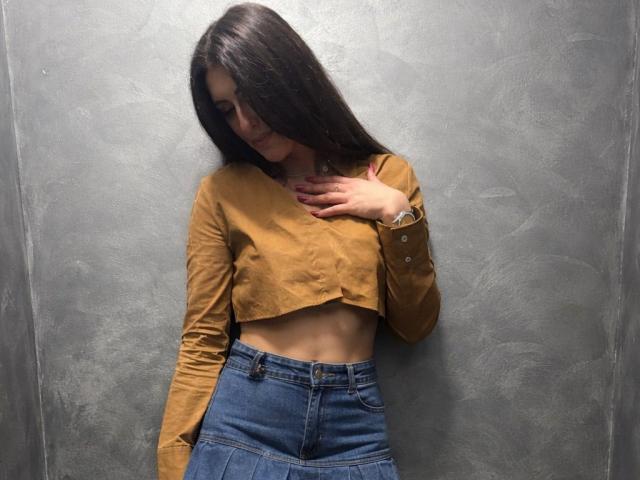 Karina Vance se tient devant un mur gris texturé, légèrement penchée vers la gauche. Elle a de longs cheveux noirs, porte un débardeur jaune moutarde et un jean bleu taille haute. Sa main effleure sa poitrine. Incroyablement sexy..