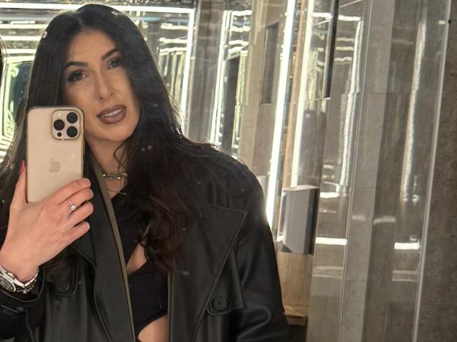 KarinaVance maakt een selfie in een moderne, helder verlichte spiegelkamer, terwijl ze met haar linkerhand een iPhone vasthoudt. Ze heeft donker, golvend haar, donkerbruine lippenstift en draagt een zwarte leren jas. Haar uitdrukking is verleidelijk..