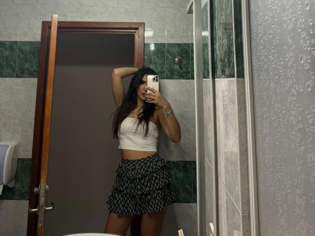 KarinaVance staat voor een spiegel in de badkamer en maakt een selfie, met haar arm omhoog, gekleed in een witte crop top en een zwarte rok met een patroon. Natte tegels en een glazen douche op de achtergrond. Een telefoon verbergt gedeeltelijk het gezicht. Heel aantrekkelijk..