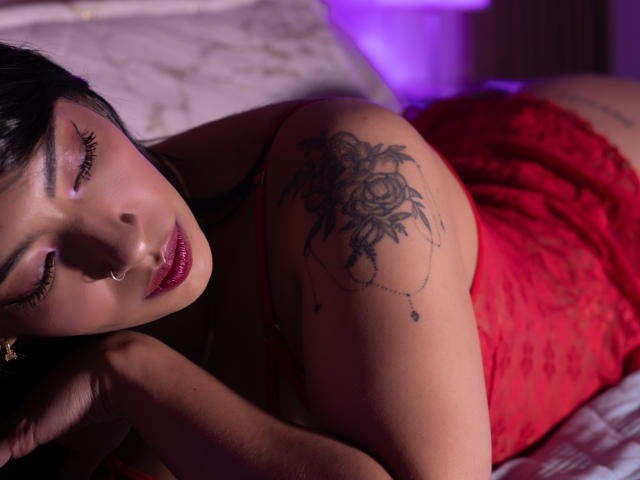ZoediMaria liegt auf der Seite, trägt ein rotes Spitzenkleid, hat die Augen halb geöffnet und glänzende, rote Lippen. Auf ihrem Körper befindet sich ein schwarzes, florales Tattoo. Schulter, gedämpftes, violettes Licht im Hintergrund, sinnlich, entspannte Pose..