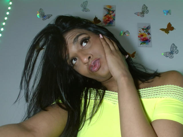 IdiiaLatina with black hair, pouty lips, and hand on cheek, wearing a yellow off-shoulder top, surrounded by colorful butterfly stickers and fairy lights, exuding sexual tension.