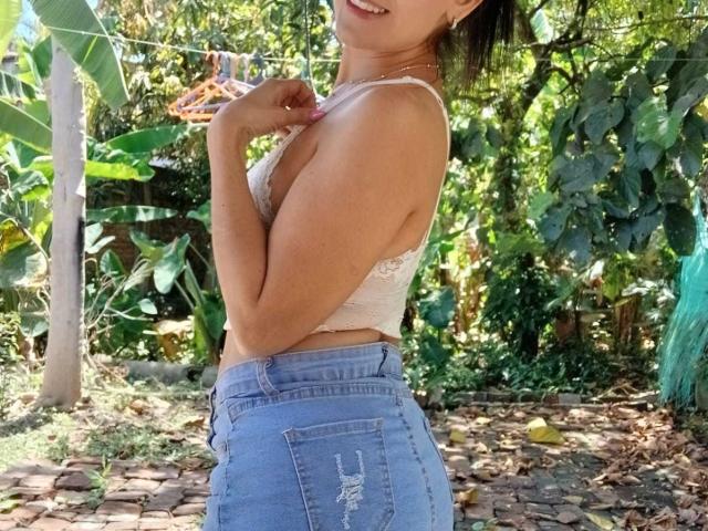 SolaraDubois, in piedi all'aperto, vista laterale, indossa un reggiseno in pizzo bianco e jeans blu a vita alta, illuminata dalla luce del sole. filtrando attraverso un lussureggiante fogliame verde, mordendosi leggermente il labbro inferiore, un accenno di tensione sessuale, posa casuale..