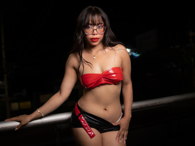 CarlaLordes, com cabelo preto molhado, sutiã de látex vermelho e shorts de couro preto com alça vermelha, em pé contra um fundo escuro. fundo, mão no corrimão, olhar intenso, batom vermelho, sexy, confiante.