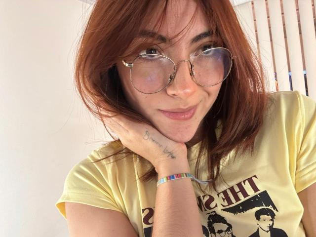 Primo piano di SpencerLe, una donna con i capelli rossi e gli occhiali, che sorride mentre appoggia la guancia sulla mano. Indossa una maglietta gialla con una scritta nera e un braccialetto color arcobaleno. Che carino!.