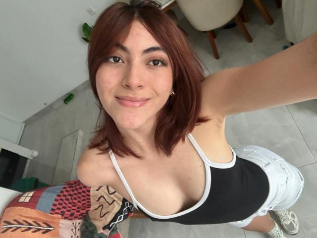 SpencerLe, com cabelo castanho na altura dos ombros, sorrindo para a câmera, vestindo uma camiseta preta e uma saia branca de renda, esticando um braço para tirar uma selfie, mostrando o colo, ambiente interno casual..