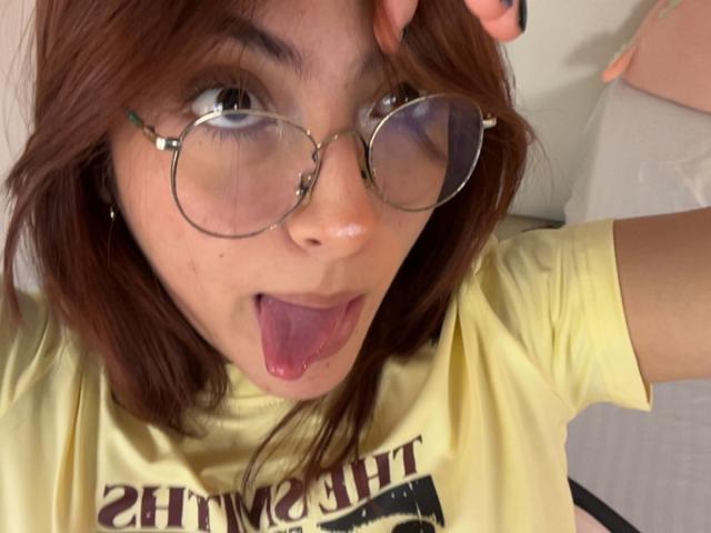 Fotografía de primer plano de SpencerLe con cabello castaño despeinado, gafas redondas y una camisa amarilla. Lengua afuera, ojos muy abiertos, mano tocando las gafas. Tensión sexual expresada de forma juguetona y provocativa. Iluminación brillante.