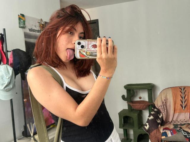 SpencerLe, com cabelo ruivo de comprimento médio, de língua para fora, segurando um telefone com capa estampada, vestindo uma regata preta. borda branca, alça verde, tirando selfie, ambiente de quarto, arranhador para gatos ao fundo..