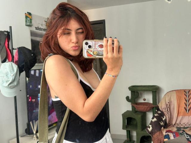 SpencerLe, con cabello castaño hasta los hombros, vistiendo una blusa negra y blanca, tomando una selfie frente al espejo con una funda para el teléfono. El fondo incluye un perchero, una estantería verde y un cojín de colores. Increíblemente atractivo/a..