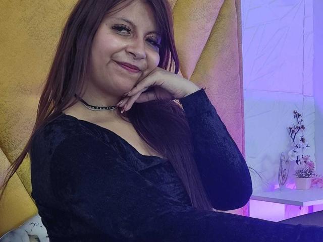 AmbeRouse with long brown hair, smiling, wearing a black lace top and choker, resting her hand on her cheek, sitting near a yellow wall and purple-lit shelf with plants.