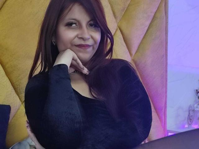 AmbeRouse with long brown hair, wearing a black top, leaning on her right hand, smiling subtly. Yellow quilted background, blue-purple light on the right. Sexual tension from her gaze.