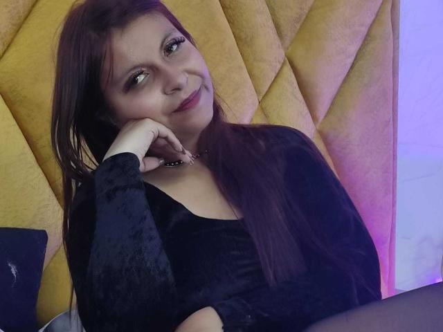 AmbeRouse with long brown hair, smiling seductively, rests her chin on her hand. Wearing a black top, yellow quilted wall in the background, soft purple lighting on the right.