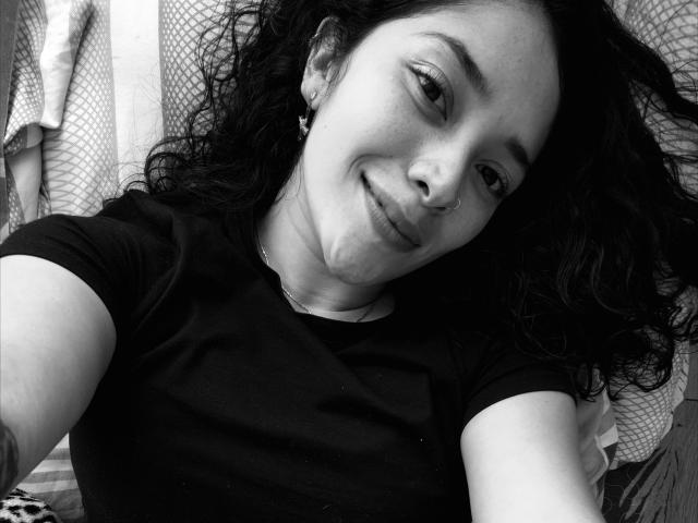 Black-and-white selfie of ChantalBernadette, lying down with messy dark hair, smiling slightly, wearing a black t-shirt. Background includes mesh and vertical stripes. High contrast, sexual tension implied.