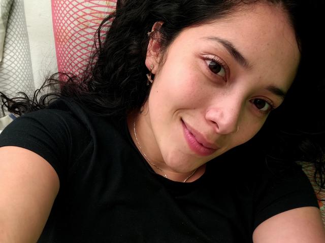 Foto em close-up de ChantalBernadette, com cabelo preto ondulado, pele clara e olhos castanhos, sorrindo levemente. Ela está vestindo uma camiseta preta e está deitada em uma superfície com um padrão vermelho e branco. O pênis dela não está visível..