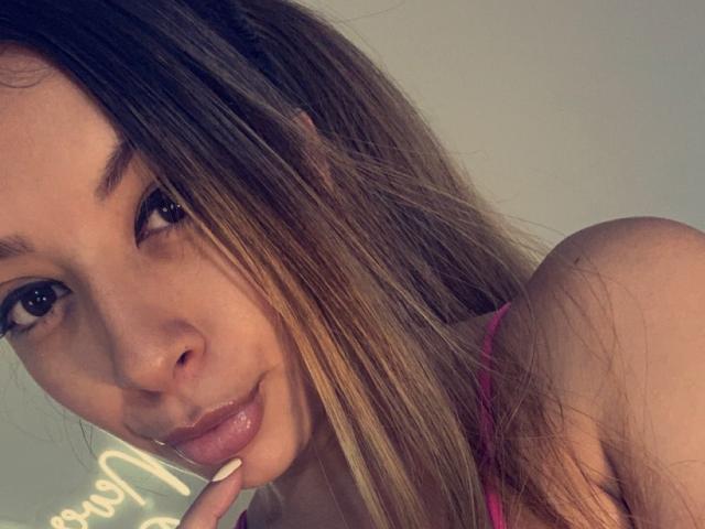 SunnySmith11 heeft glanzende bruine ogen, een lichte huid en steil bruin haar met blonde highlights. Ze draagt een roze top en heeft een vinger op haar licht glanzende lippen..