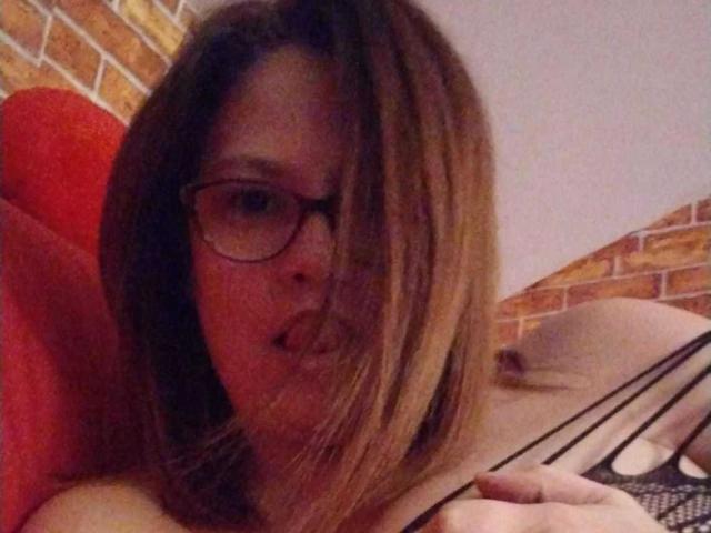 Close-up photo of HellenBooty69, a young woman with glasses and shoulder-length brown hair, sticking her tongue out while leaning against a red pillow and black fishnet fabric. Sexual tension is palpable.