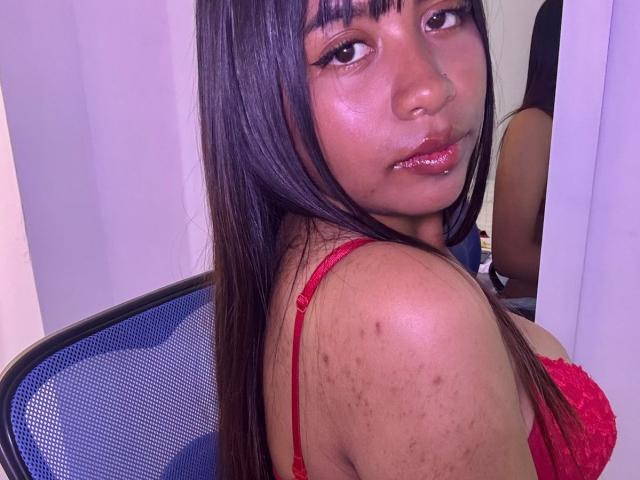 Close-up of NahomyNoir, a young woman with long black hair, wearing a red lace bra, looking over her shoulder. She has dark brown skin with acne marks on her shoulder. Blue chair in background.