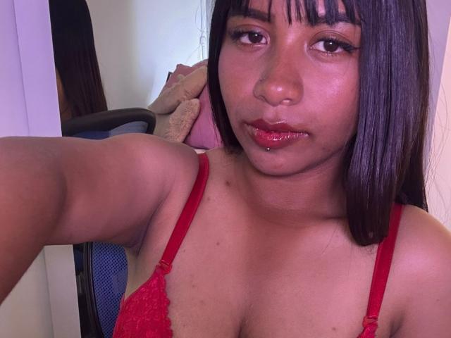 Photo en gros plan de NahomyNoir, avec une peau marron moyen, des cheveux noirs lisses avec une frange, portant un soutien-gorge en dentelle rouge. Elle lèche ses lèvres rouges et brillantes, une main dans ses cheveux, une tension sexuelle palpable..