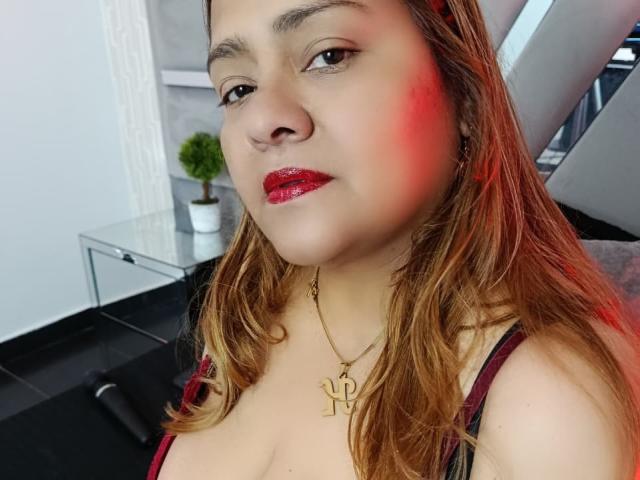 Primer plano de Monzerrat Boyer, de piel clara con rubor rojo en la mejilla, que lleva lápiz labial rojo y un collar dorado con una "L". collar. Cabello castaño, camiseta negra sin mangas, expresión sensual, iluminación roja tenue..