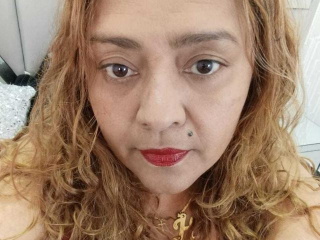 Close-up foto van Monzerrat Boyer, een vrouw van middelbare leeftijd met krullend rood haar, rode lippenstift en een piercing in haar gezicht. linkerkwang.

ze draagt een gouden ketting met een 'P'-bedeltje. Verdomd sexy uitdrukking..