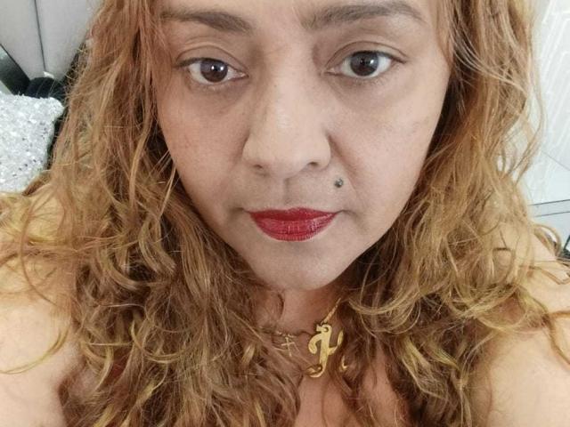 Close-up foto van Monzerrat Boyer, een vrouw van middelbare leeftijd met krullend rood haar, rode lippenstift en een klein beauty markje op haar gezicht. linkerkwang.

ze draagt een gouden ketting en kijkt recht in de camera met een lichte grijns. Haar ogen zijn bruin en ze heeft een sexy, zelfverzekerde uitdrukking..