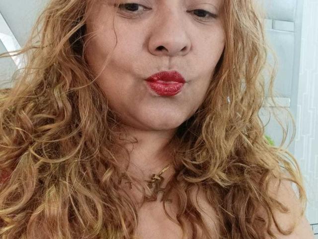 Close-up of MonzerratBoyer with curly blonde hair, red lipstick, and a gold necklace, puckering her lips suggestively. Her eyes are slightly squinted, creating a seductive expression. Focused on her face.