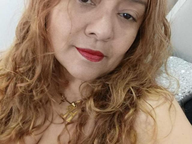 Close-up foto van Monzerrat Boyer, een witte vrouw met golvend, roodbruin haar, rode lippenstift en een gouden ketting. Ze heeft een lichte glimlach, waardoor een vleugje seksuele spanning doorschemert. Haar gezicht is in focus..