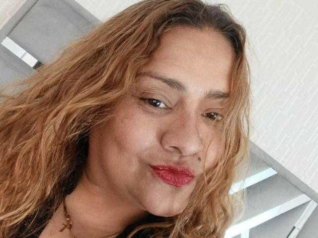 Close-up foto van Monzerrat Boyer, een vrouw met golvend, lichtbruin haar en rode lippenstift, die lichtjes naar beneden kijkt. een subtiele grijns. Hel licht in de achtergrond. Seksuele spanning in haar blik..