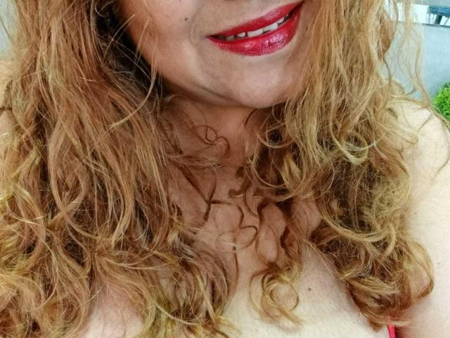 Primer plano de Monzerrat Boyer con cabello rizado y pelirrojo, lápiz labial rojo y un ligero escote. Sus labios brillantes y su boca ligeramente abierta crean una atmósfera sugestiva y erótica. La iluminación brillante resalta sus rasgos..