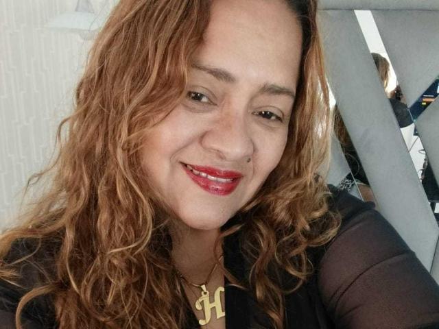 Close-up de Monzerrat Boyer, sorrindo com batom vermelho, cabelo castanho ondulado, blusa preta, colar dourado com um pingente em formato de "H". Assento de carro visível e janela ao fundo, tensão sexual, rosto atraente..