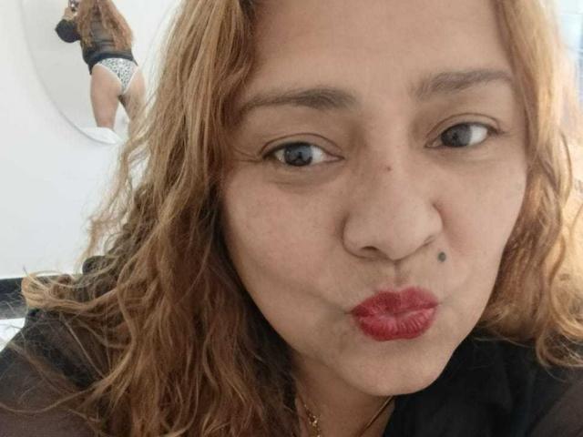 Foto em close-up de Monzerrat Boyer, de pele clara, com cabelo loiro ondulado, lábios vermelhos franzidos, olhos azuis e uma pequena pinta acima. lábio esquerdo, parte superior preta, joias discretas, luz suave, leve sombra, pose casual.