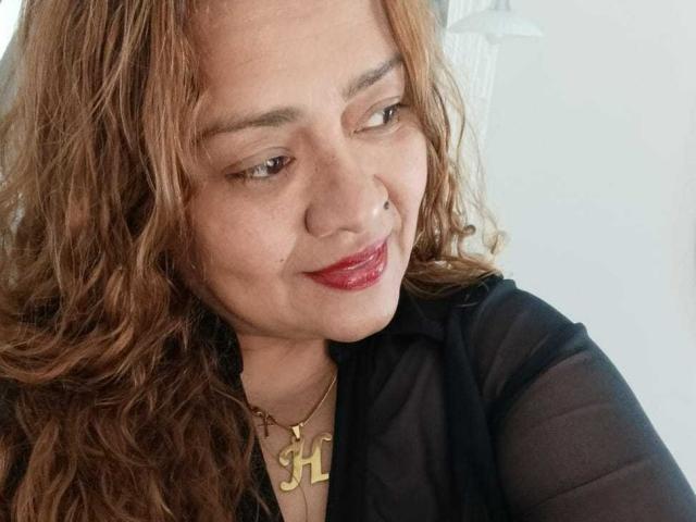 Close-up de Monzerrat Boyer, uma mulher de meia-idade com cabelo castanho ondulado, batom vermelho e um piercing no nariz. Ela veste uma blusa preta e um colar dourado com a letra "H". Pequeno sorriso, tensão sensual..