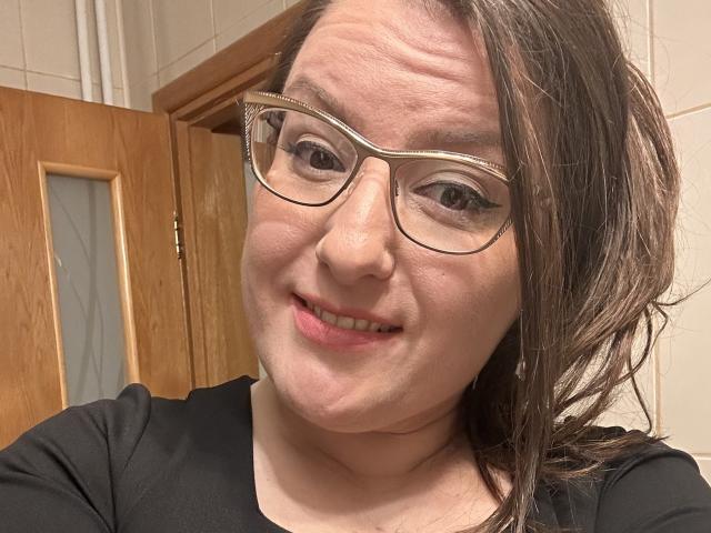 Gros plan de BadAngel, avec une peau claire, des cheveux bruns et des lunettes, souriant légèrement. BadAngel porte une chemise noire, et se trouve devant des portes en bois. Une légère tension sexuelle dans le regard..