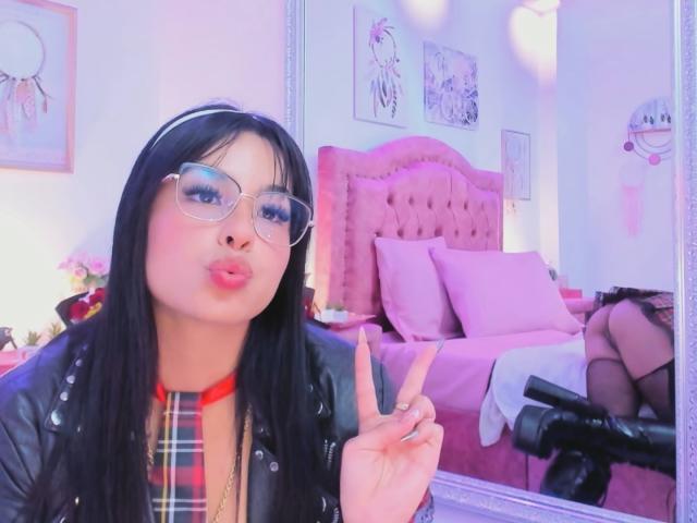 Photograph of GraceClarke with long black hair, wearing glasses and a black leather jacket, making a peace sign, pouting lips, plaid shirt, pink bed in the background, legs in stockings.