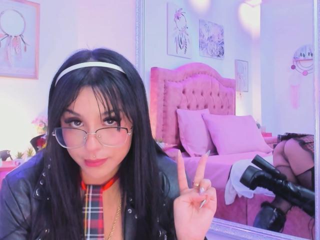 Photograph of GraceClarke with long black hair, wearing glasses, white headband, black leather jacket, plaid shirt, flashing a middle finger, white gloves on a pink bed in a brightly lit room.