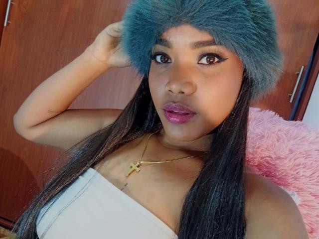 Primer plano de Caylin con un sombrero de piel gris, un top blanco sin tirantes y lápiz labial púrpura. Ella sostiene el sombrero con una mano, revelando un collar con una cruz de oro. Almohada rosa y esponjosa en el fondo. Increíblemente sexy..