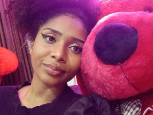 Megan Williams, una mujer de piel oscura con cabello afro natural, delineador de ojos negro y labios brillantes, se inclina cerca de un gran peluche rojo. Animal con un hocico negro. Tensión sexual, mirada directa, pose íntima.