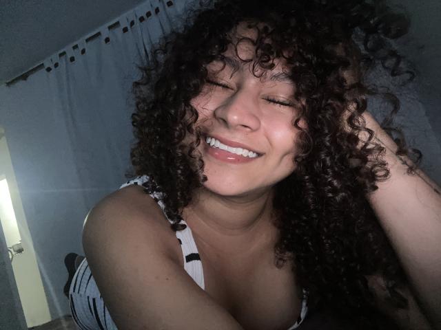 Photograph of SofiaPassionne with dark curly hair, smiling widely, eyes closed, hand in hair, wearing a white tank top with black stripes, showing cleavage, dimly lit room, curtains in background.