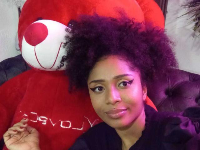 Photographie de Megan Williams, avec des cheveux noirs et bouclés, assise sur un canapé en cuir noir, appuyée contre un grand ours en peluche rouge. Un ours avec un visage blanc et des yeux rouges en forme de boutons. Son expression est neutre..