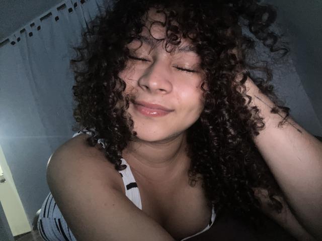 Fotografia de SofiaPassionne com cabelo cacheado escuro, olhos fechados, sorrindo levemente, vestindo uma regata listrada, com a mão em cabelo, quarto com pouca luz, fundo sombrio, luz suave no rosto, clima sensual..