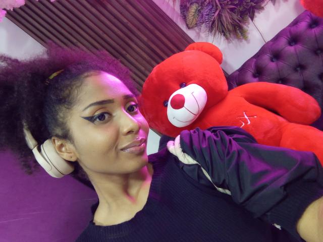 Megan Williams, avec une peau brun clair et des cheveux bouclés, portant une chemise noire, tenant un grand ours en peluche rouge avec un nez blanc. et sourire, avec des écouteurs blancs, éclairage violet, sourire séduisant, pose décontractée..