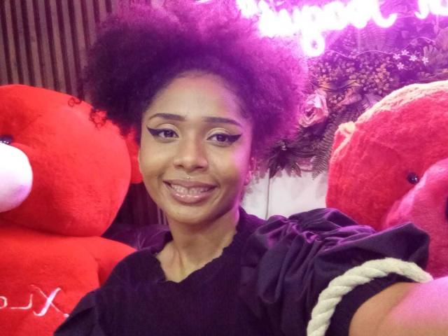 Megan Williams, con un'ampia acconciatura afro, sorride con occhi scintillanti, indossa una maglietta nera ed è circondata da due grandi peluche rossi. cuori, con un bastone per selfie, illuminazione vibrante in tonalità di viola e rosa.