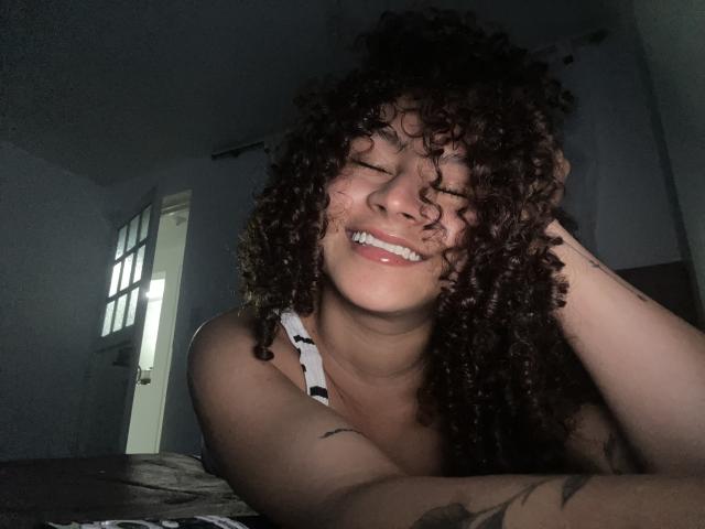 Fotografia di SofiaPassionne con capelli ricci scuri, che sorride mostrando i denti, con una mano tra i capelli, pelle marrone chiaro, vestita di nero. canotta, tatuaggio sul braccio, stanza poco illuminata, finestra sullo sfondo. Molto attraente..