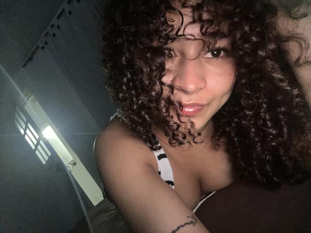 Close-up of SofiaPassionne, a dark-skinned woman with curly black hair, wearing a black and white striped top, leaning forward with a seductive smile. Bright light on the left side.