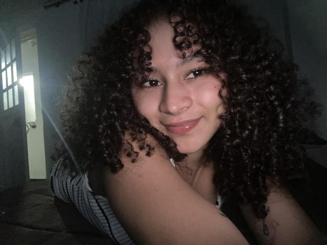 Fotografía de SofiaPassionne con cabello negro, rizado y abundante, sonriendo ligeramente. Tiene un tono de piel marrón claro, ojos marrones y un piercing en la nariz. La luz resalta su rostro, creando un brillo sensual..