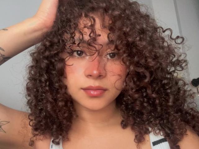 Close-up de SofiaPassionne com cabelo cacheado escuro e desalinhado, mão no cabelo, olhos castanhos, lábios cheios, pele marrom clara, tatuagens. em um braço, vestindo uma camiseta branca, sexy, olhar intenso, luz natural..