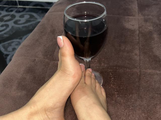 Twee blote voeten, met netjes geknipte nagels, waarbij één voet op de andere rust, raken een glas donkerrode wijn dat op een bruine ondergrond staat. Gestructureerd oppervlak.
heerlijk ontspannen, intieme sfeer..