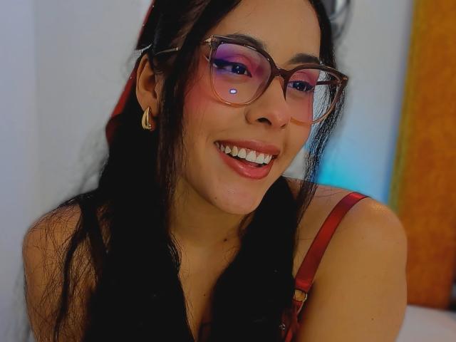 Gros plan de MarilynHills, souriante, portant des lunettes à monture rose, des cheveux noirs, une robe rouge avec des bretelles, un boucle d'oreille doré et un nez visible. perçant, légèrement rougi, lumière vive, fond bleu doux, ambiance sexy.